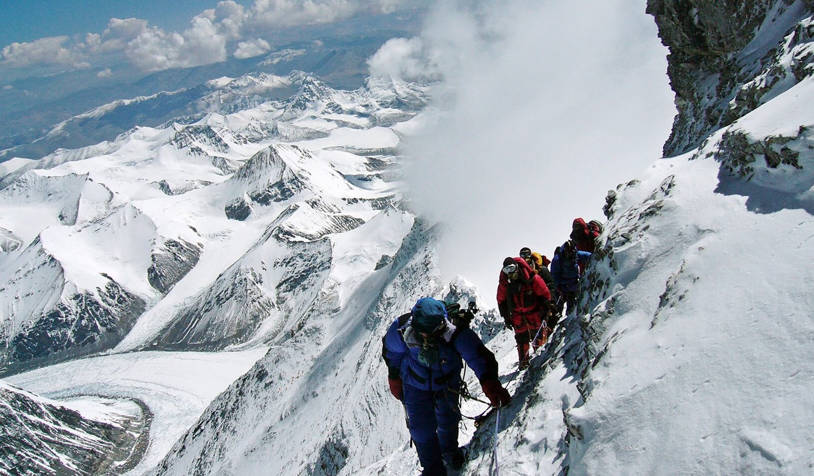 A rope team climbing a snowy summit ridge high above a glacier